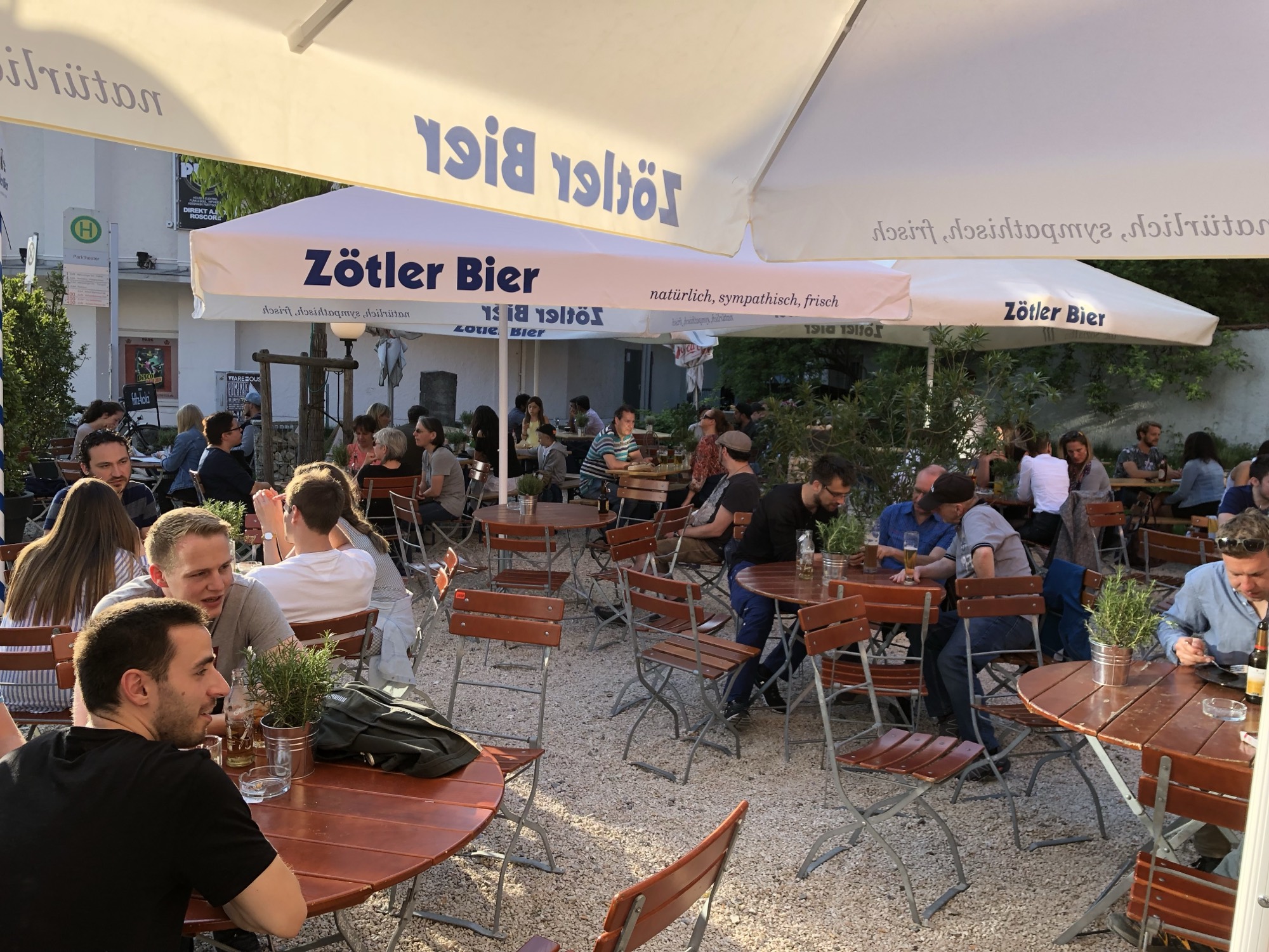Voller Biergarten des Künstlerhauses an einem Sommerabend – Kiesboden, Klappstühle, Zötler-Schirme
